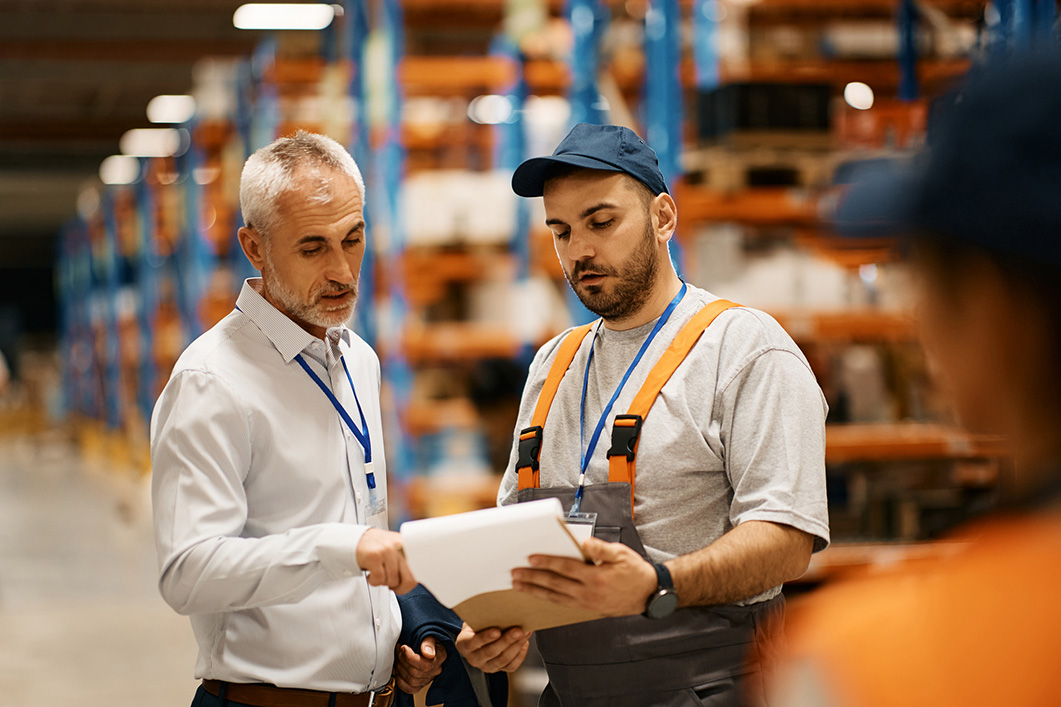 Mature inspector and warehouse worker cooperating while going through paperwork at distribution warehouse.