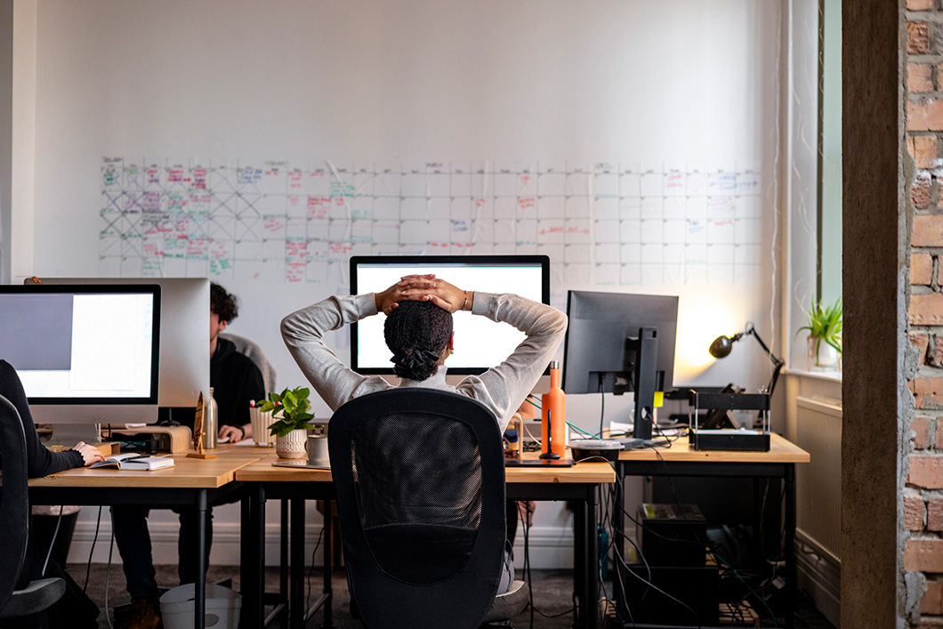 Rear view wide shot of a woman sitting working at a computer in an office in England. She has her hands behind her head as she stretches, looking at the screen.
