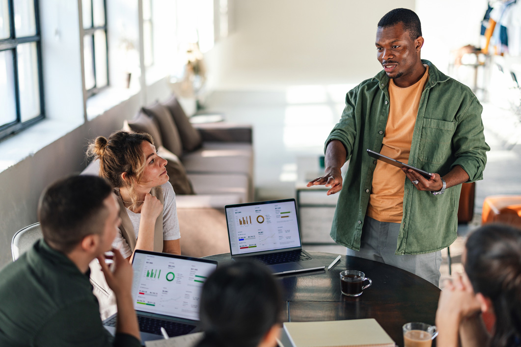 A group of diverse young professionals engaged in a collaborative discussion in a modern office setting, utilizing technology for effective teamwork and project management.