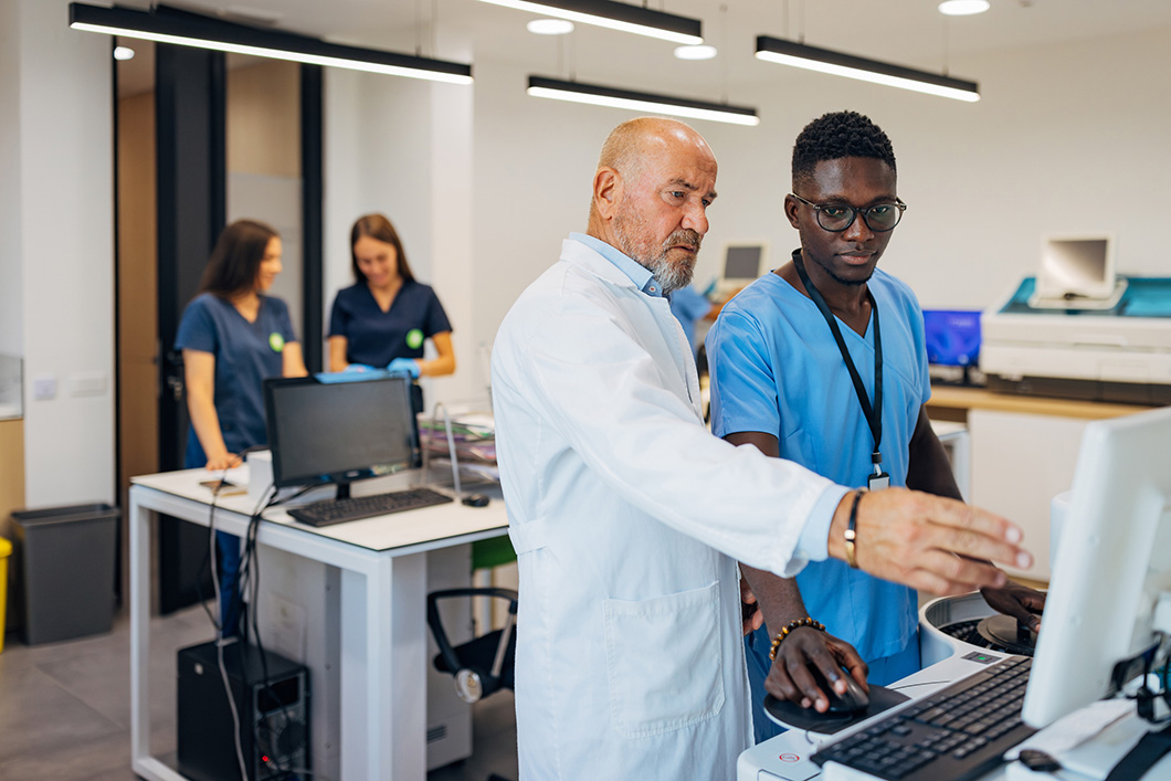 A senior male scientist in a lab coat shows a student on the computer the results of medical tests performed in the laboratory
