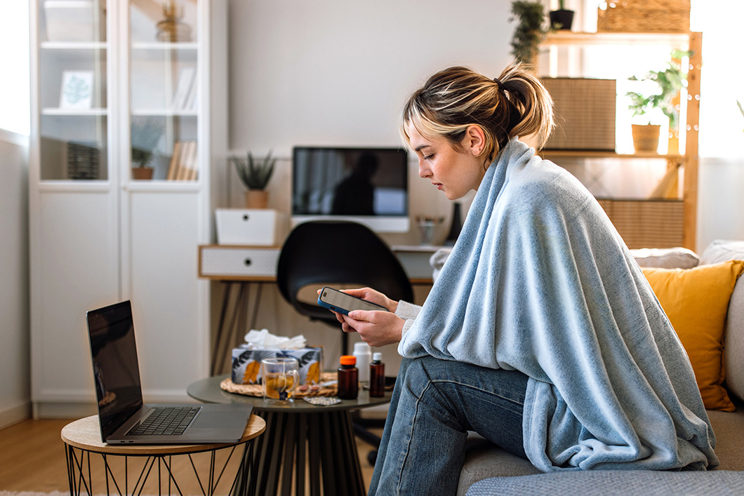 An employee is working from home, at a laptop, with a blanket wrapped around her and some medicine.